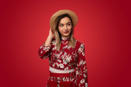Photo for Self assured young female model with long blond hair in floral dress adjusting straw hat and looking away against red background - Royalty Free Image
