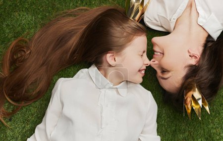 Photo for From above attractive adult woman and adorable girl in paper crowns smiling and touching noses while lying on green grass together - Royalty Free Image