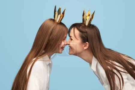Téléchargez les photos : Vue latérale de la petite fille et de la femme portant des couronnes d'or et faisant grimace les uns aux autres debout en face sur fond bleu - en image libre de droit