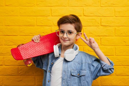 Téléchargez les photos : Garçon confiant à la mode en jean veste et écouteurs tenant longboard rouge signe V gestuelle et regardant la caméra contre le mur de briques jaunes - en image libre de droit