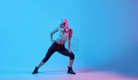 Foto de Cuerpo completo joven delgado hembra en ropa deportiva y peluca realizando embestidas y mirando a la cámara de entrenamiento blanco bajo luz de neón - Imagen libre de derechos