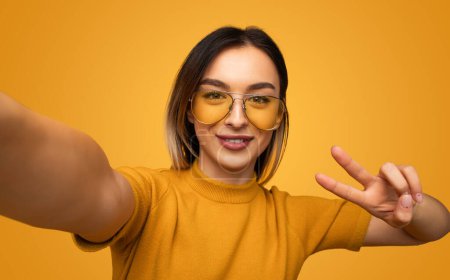 Téléchargez les photos : Femme optimiste dans des lunettes jaunes montrant le signe de victoire tout en regardant la caméra et en prenant selfie - en image libre de droit
