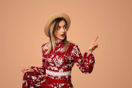 Photo for Confident young female model with blond hair in floral dress and straw hat pointing aside and looking away against beige background - Royalty Free Image