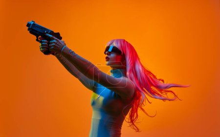 Téléchargez les photos : Vue latérale de la jeune femme en tenue futuriste et des lunettes avec de longs cheveux roses volants visant pistolet loin sur fond orange - en image libre de droit