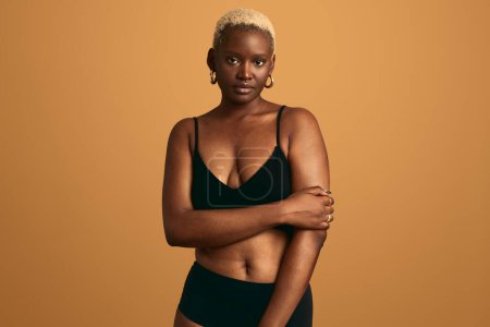 Photo for Confident African female with short blond Afro hair in black lingerie looking at camera while standing against beige background - Royalty Free Image