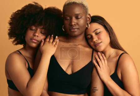 Foto de Tranquil modelos femeninos multirraciales en lencería negra abrazándose y cerrando los ojos mientras están de pie juntos sobre fondo beige - Imagen libre de derechos