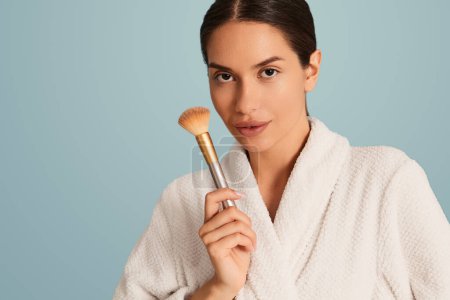 Photo for Crop confident young female model with perfect skin and dark hair in white bathrobe demonstrating makeup brush and looking at camera against blue background - Royalty Free Image