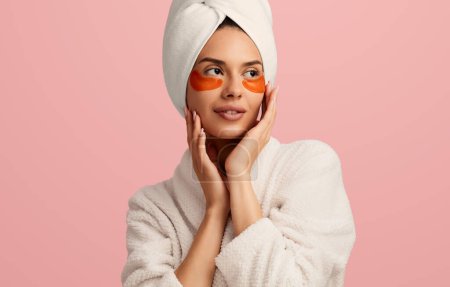 Téléchargez les photos : Jeune femme en peignoir et serviette sur la tête appliquant des patchs hydratants sur le visage et, en regardant l'espace de copie pendant la routine de soins de la peau sur fond rose - en image libre de droit