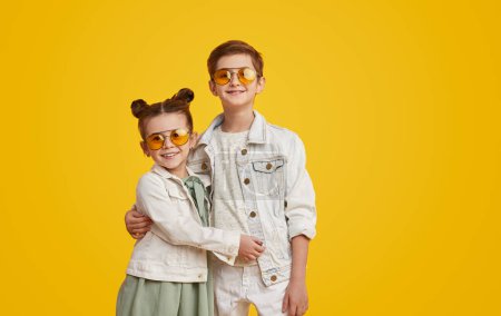 Téléchargez les photos : Garçon mignon et fille dans des vêtements modernes à la mode embrassant et regardant la caméra tout en se tenant debout sur un fond jaune vif - en image libre de droit