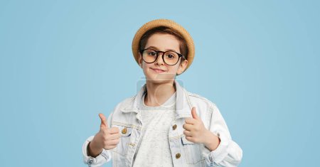 Foto de Elegante niño gafas inestables y sombrero de paja mirando a la cámara contra el fondo azul que muestra los pulgares hacia arriba gesto - Imagen libre de derechos