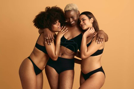 Photo for Young smiling multiethnic ladies in black lingerie hugging each other in studio against orange background support and acceptance concept - Royalty Free Image