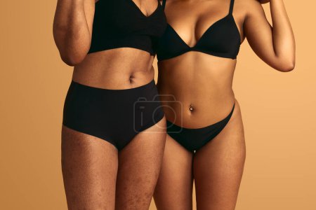 Foto de Mujeres diversas sin rostro en lencería negra mostrando positividad corporal mientras están de pie cerca sobre fondo marrón - Imagen libre de derechos