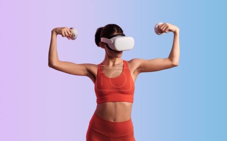 Photo for Fit muscular female athlete in red activewear and VR headset with controllers showing biceps against gradient background - Royalty Free Image