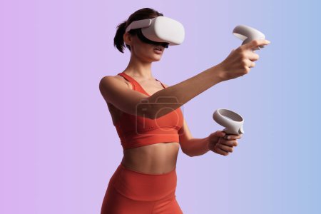 Photo for Fit young sportswoman in red sportswear and VR headset using controllers while playing virtual game against gradient background - Royalty Free Image