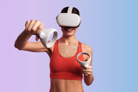 Photo for Young smiling fit woman in VR headset using controllers while playing videogame making virtual punch against gradient background - Royalty Free Image