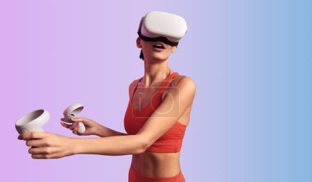 Foto de Mujer joven en camiseta deportiva roja con auriculares VR mientras juega a videojuegos utilizando controladores que exploran el ciberespacio contra el fondo degradado - Imagen libre de derechos