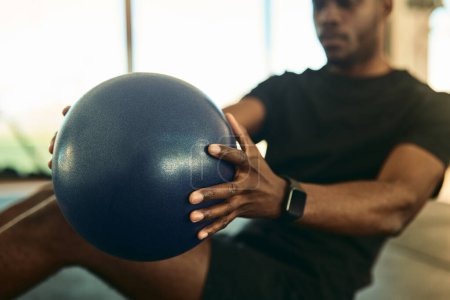 Crop concentrated young African American male athlete in activewear and fitness tracker doing abs exercise with medicine ball during workout in gym