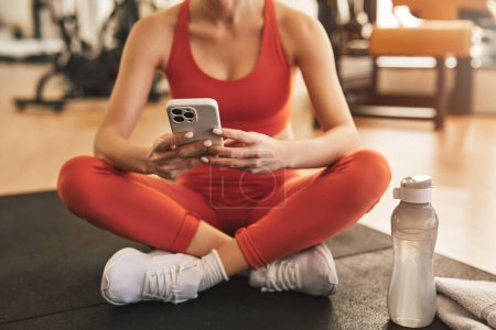Photo for Crop anonymous female in activewear sitting on floor in lotus pose and browsing mobile phone while resting after workout in gym - Royalty Free Image