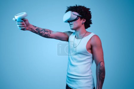 Foto de Vista lateral de un joven hispano joven y serio millennial con aire rizado oscuro y tatuajes, pulsando el botón en el controlador mientras juega a videojuegos en gafas VR en estudio azul - Imagen libre de derechos