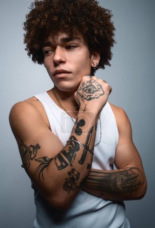 Foto de Confiado joven modelo masculino hispano serio con tatuajes en los brazos y pelo oscuro rizado en la parte superior blanca, tocando la cara y mirando hacia otro lado contra el fondo gris en el estudio - Imagen libre de derechos