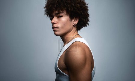 Photo for Side view of serious self assured young fit Hispanic male model with curly dark hair in white top gazing at camera against gray background - Royalty Free Image
