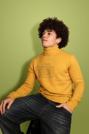 Foto de Modelo masculino étnico joven y seguro de sí mismo con cabello afro oscuro en cuello alto amarillo con estilo, sentado en el taburete con la mano en el bolsillo y mirando hacia otro lado contra el fondo verde - Imagen libre de derechos