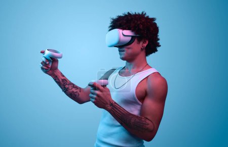 Photo for Serious male in VR goggles and with controllers playing video game against blue background - Royalty Free Image