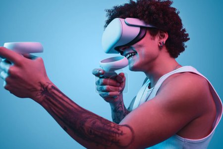 Photo for Side view of young African American male with afro hairstyle in VR goggles interacting with virtual reality against blue background - Royalty Free Image