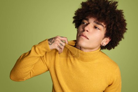 Foto de Joven hombre hispano con peinado afro en camisa de cuello alto amarilla mirando hacia otro lado sobre fondo verde - Imagen libre de derechos