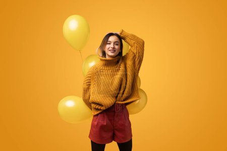 Foto de Jovencita moderna en traje elegante sosteniendo racimo de globos vivos mientras está de pie contra el fondo amarillo - Imagen libre de derechos