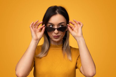 Trendy female in bright outfit and adjusting stylish shades and looking to the side against yellow background