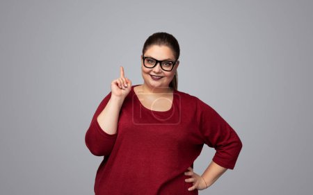 Photo for Content smart plus size woman with long dark ponytail and red lips in eyeglasses, smiling and looking at camera while pointing up standing against gray background with hand on waist - Royalty Free Image