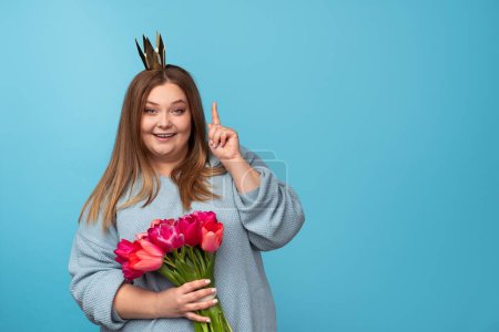 Foto de Encantadora hembra regordeta con ramo de hermosos tulipanes y corona de papel sonriendo y mirando a la cámara mientras señala hacia arriba sobre fondo azul - Imagen libre de derechos