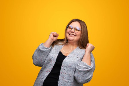 Photo for Happy young plump female in gray cardigan and eyeglasses smiling while clenching fists and rejoicing over achievement against yellow background - Royalty Free Image