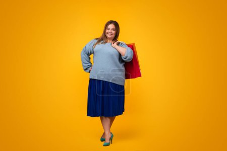 Foto de Bastante más tamaño femenino en traje de moda sonriendo y sosteniendo bolsas de papel mientras está de pie sobre fondo amarillo - Imagen libre de derechos