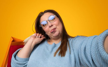Foto de Mujer regordeta asombrada en gafas con estilo sosteniendo bolsas de papel y haciendo expresión divertida mientras toma selfie sobre fondo amarillo - Imagen libre de derechos