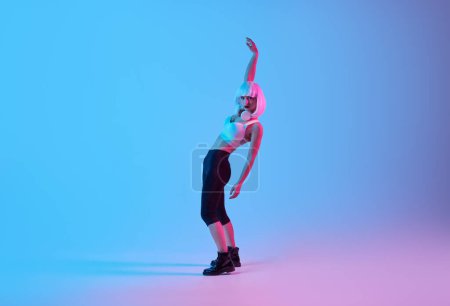 Photo for Full body side view of futuristic fashion young woman in neon light dancing with raised arms on pink background in studio - Royalty Free Image
