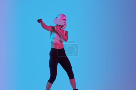 Foto de Joven hembra en ropa deportiva y auriculares VR perforando objetos invisibles mientras juega videojuego contra fondo azul - Imagen libre de derechos