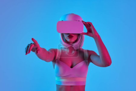 Photo for Young futuristic female in VR goggles interacting with virtual reality against blue background in neon light - Royalty Free Image