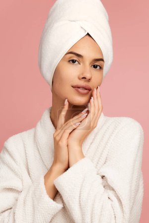 Photo for Positive young female model with perfect skin in white bathrobe and towel on head touching face and looking at camera against pink background - Royalty Free Image