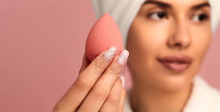 Photo for Soft focus of young woman in white bathrobe with towel on head showing makeup sponge to camera while looking away against pink background - Royalty Free Image