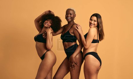 Foto de Vista lateral de felices tres mujeres multirraciales usando ropa interior negra y mostrando positividad corporal en el estudio - Imagen libre de derechos