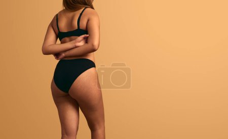 Foto de Vista posterior de una mujer anónima en lencería negra de pie con brazos cruzados sobre fondo beige con espacio para copiar - Imagen libre de derechos