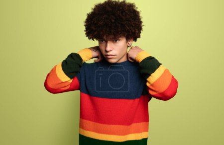Foto de Modelo masculino hispano joven serio y seguro de sí mismo con cabello rizado oscuro en suéter a rayas de colores ajustando el cuello y mirando hacia otro lado contra el fondo verde - Imagen libre de derechos