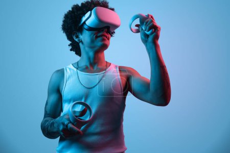 Téléchargez les photos : Jeune joueur masculin ethnique souriant aux cheveux afro dans un casque VR, explorant le cyberespace tout en jouant au jeu vidéo à l'aide de contrôleurs en studio éclairés par la lumière bleue néon - en image libre de droit