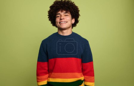 Foto de Joven positivo hispano milenial masculino con pelo rizado afro oscuro en suéter a rayas de colores, sonriendo y mirando a la cámara mientras está de pie sobre fondo verde en el estudio - Imagen libre de derechos