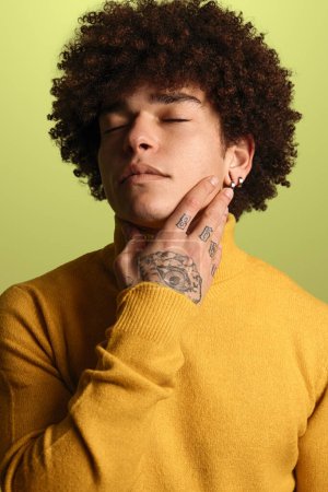 Foto de Calma joven hispano masculino millennial con pelo rizado oscuro y tatuaje en la mano en cuello alto amarillo tocando cuello con los ojos cerrados sobre fondo verde - Imagen libre de derechos