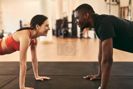 Photo for Side view of smiling African American male personal coach doing plank exercise with female athlete in sportswear on mat during intense workout at gym - Royalty Free Image