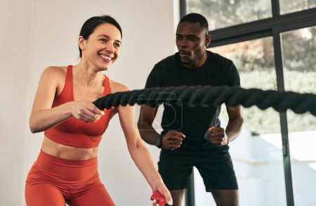 Photo for Positive fit female athlete in sportswear training in gym with battle ropes and ethnic friend is cheering and motivating - Royalty Free Image