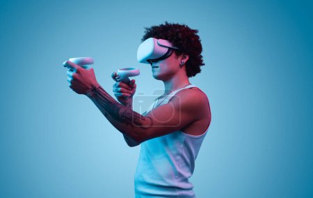 Téléchargez les photos : Vue latérale du jeune homme hispanique avec tatouage tout en se tenant debout dans des lunettes VR et avec des contrôleurs, jouer à un jeu vidéo et profiter du cybermonde sur fond bleu - en image libre de droit